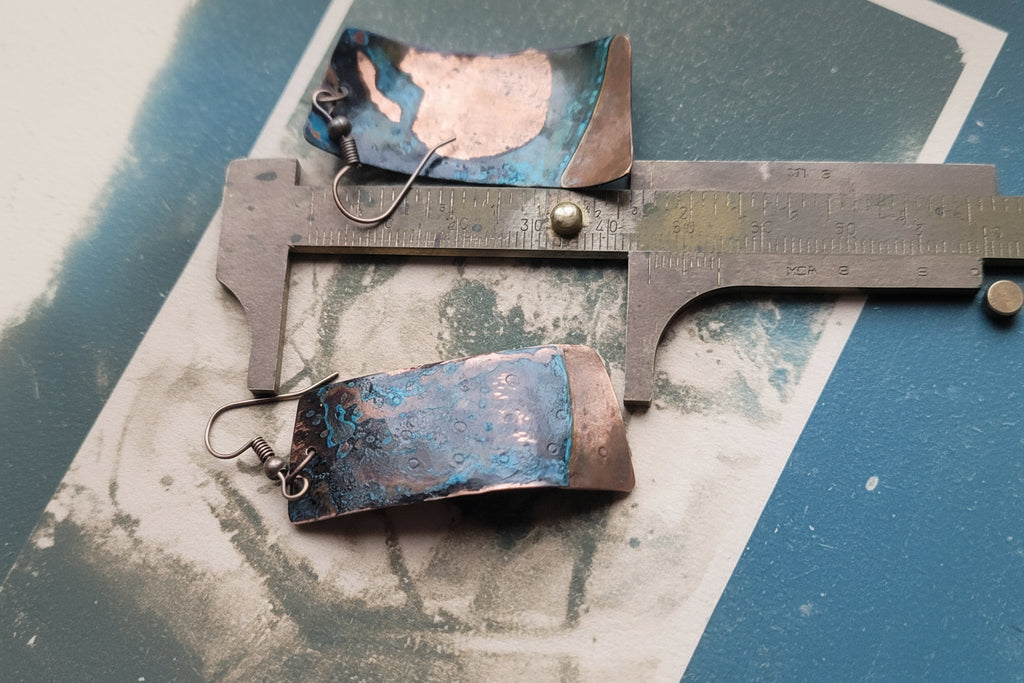 Pair of earrings with blue and brown design on a metal surface with a ruler for scale.