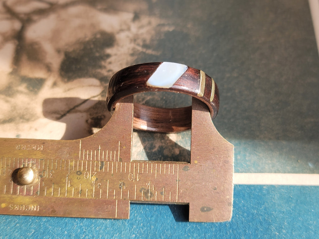 Wooden ring with brass and mother of pear being measured by a vernier caliper against a blue background.