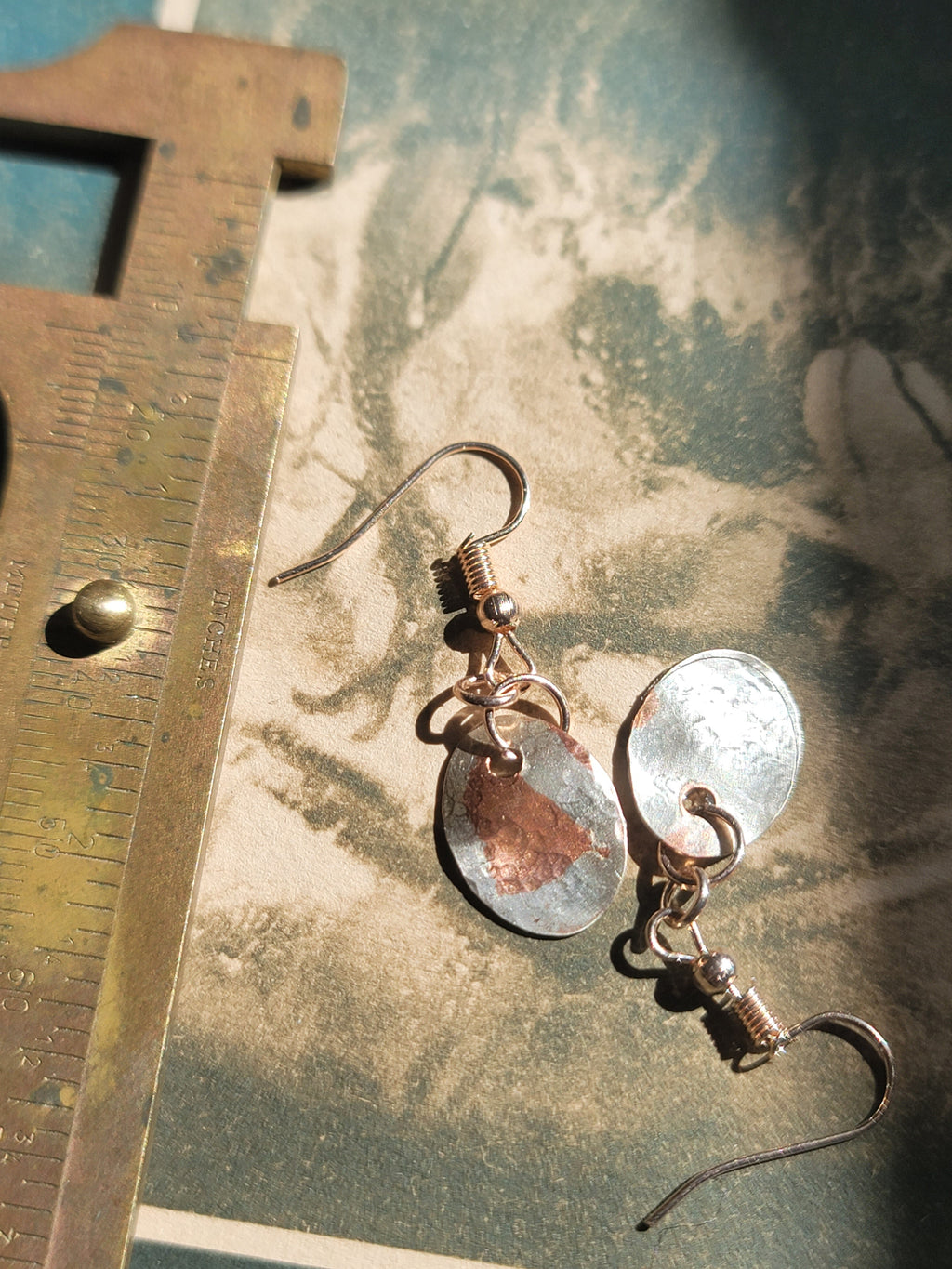 Pair of earrings with metallic components on a textured surface with a ruler for scale.